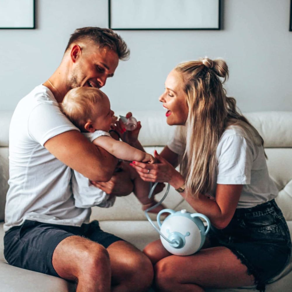 Mama i tata wspólnie oczyszczają nosek dziecka za pomocą elektrycznego aspiratora do nosa Nosiboo Pro
